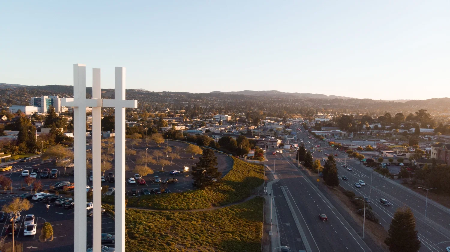 Aerial view of a cityscape featuring three large white crosses, with a highway and parking lot below, surrounded by hills.