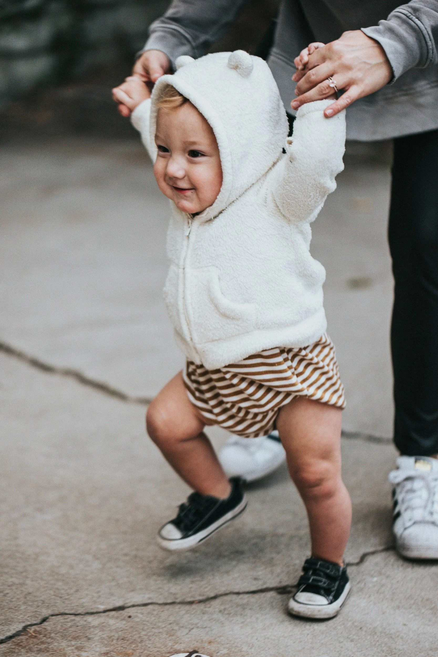 baby in hooded jacket taking steps while adult holds their hands