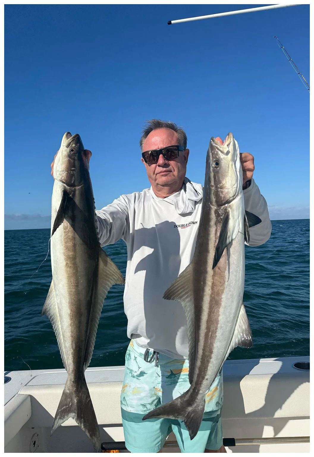 A man on a boat holding two large fish against a blue ocean and sky.
