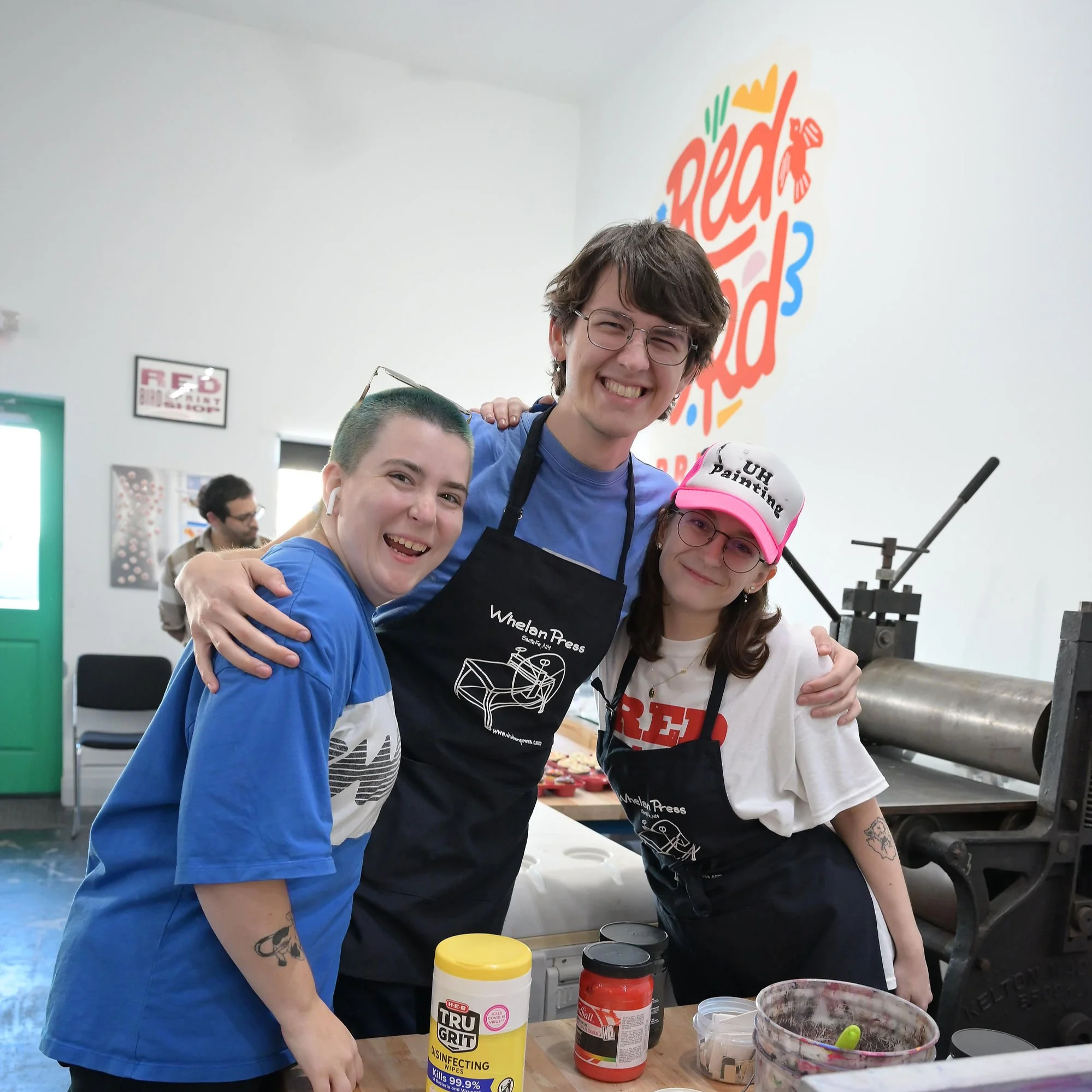 Three people smiling and hugging in a kitchen or workshop with cooking or printing equipment. They are wearing aprons with 'Whelan Press' on them. A large colorful 'Red Food 3' sign is on the wall behind them.