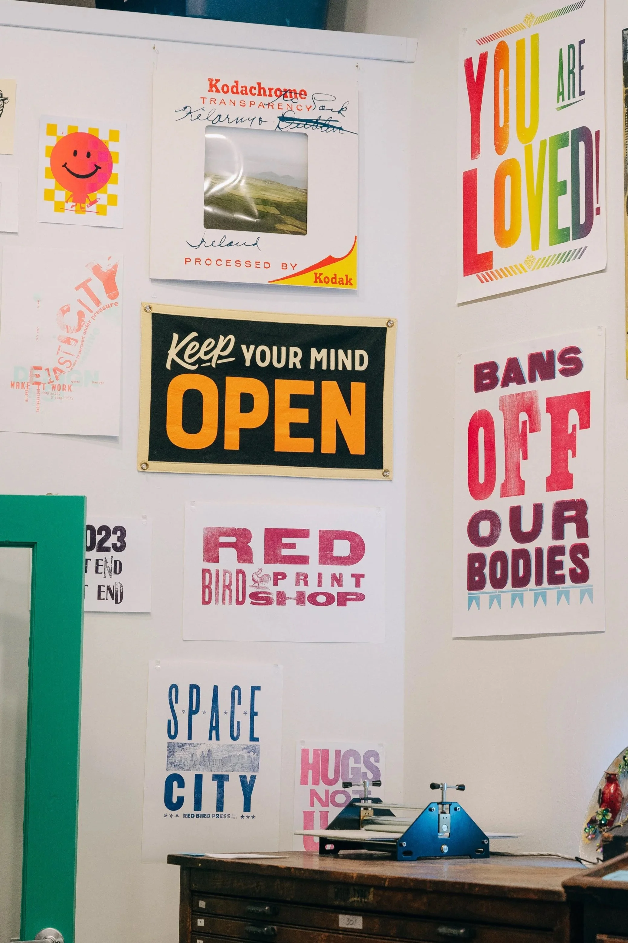 Wall with various colorful posters and prints, including phrases like "You Are Loved!", "Keep Your Mind Open", "Bans Off Our Bodies", "Space City", and "Hugs Not Hate." There is a wooden cabinet below the posters, a green door frame on the left, and a small stack of black trays or disks on top of the cabinet.