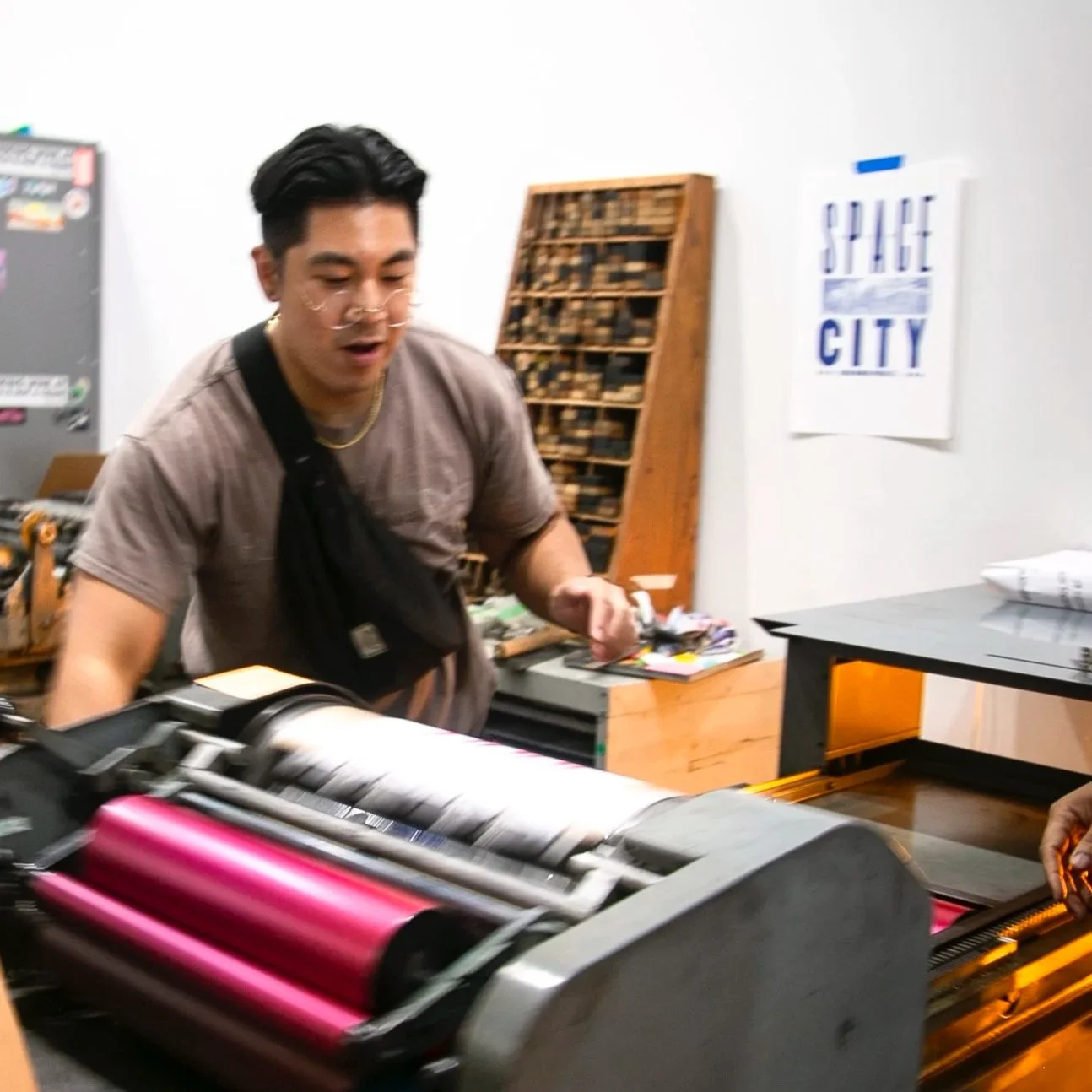 A man operating a printing press machine with pink and white rollers in a print shop, with a 'SPACE CITY' poster on the wall.