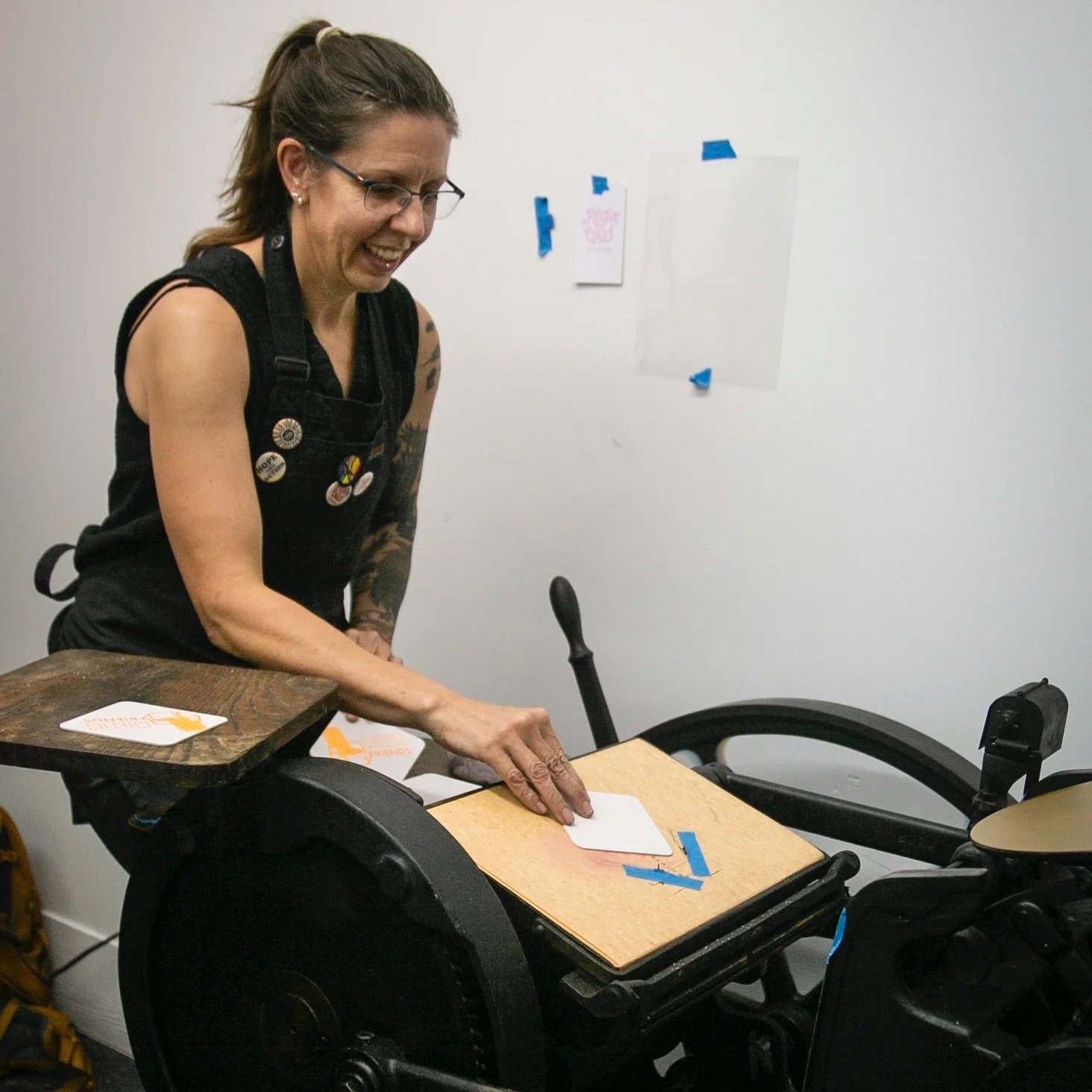 A woman with glasses and tattoos, smiling, places a card on a wooden board attached to the armrest of a wheelchair.
