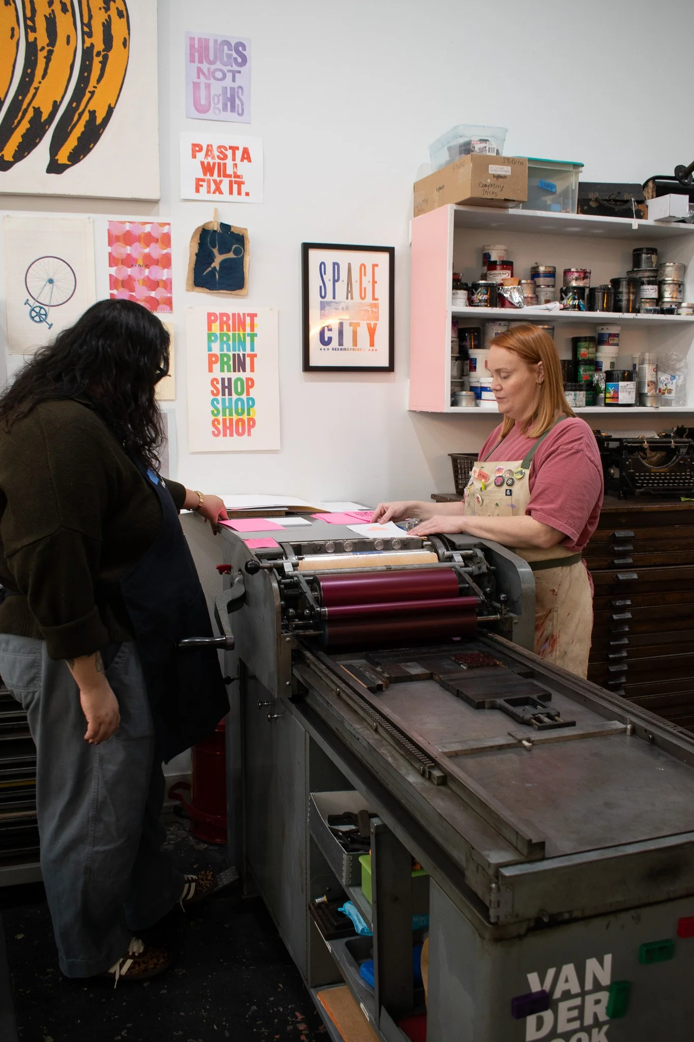 Two women working in a print shop, one operating a printing machine and the other observing, with colorful wall art and containers of ink in the background.