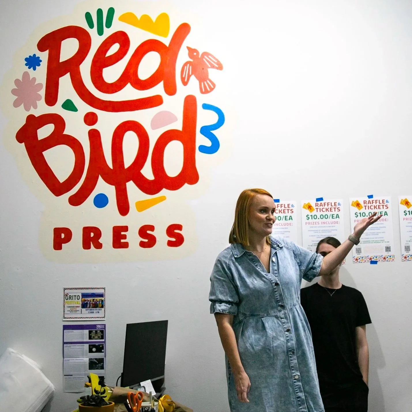 A woman at a raffle press event with a large sign that reads 'Red Bird PRESS' on the wall behind her, and posters advertising raffle tickets for prize drawings.