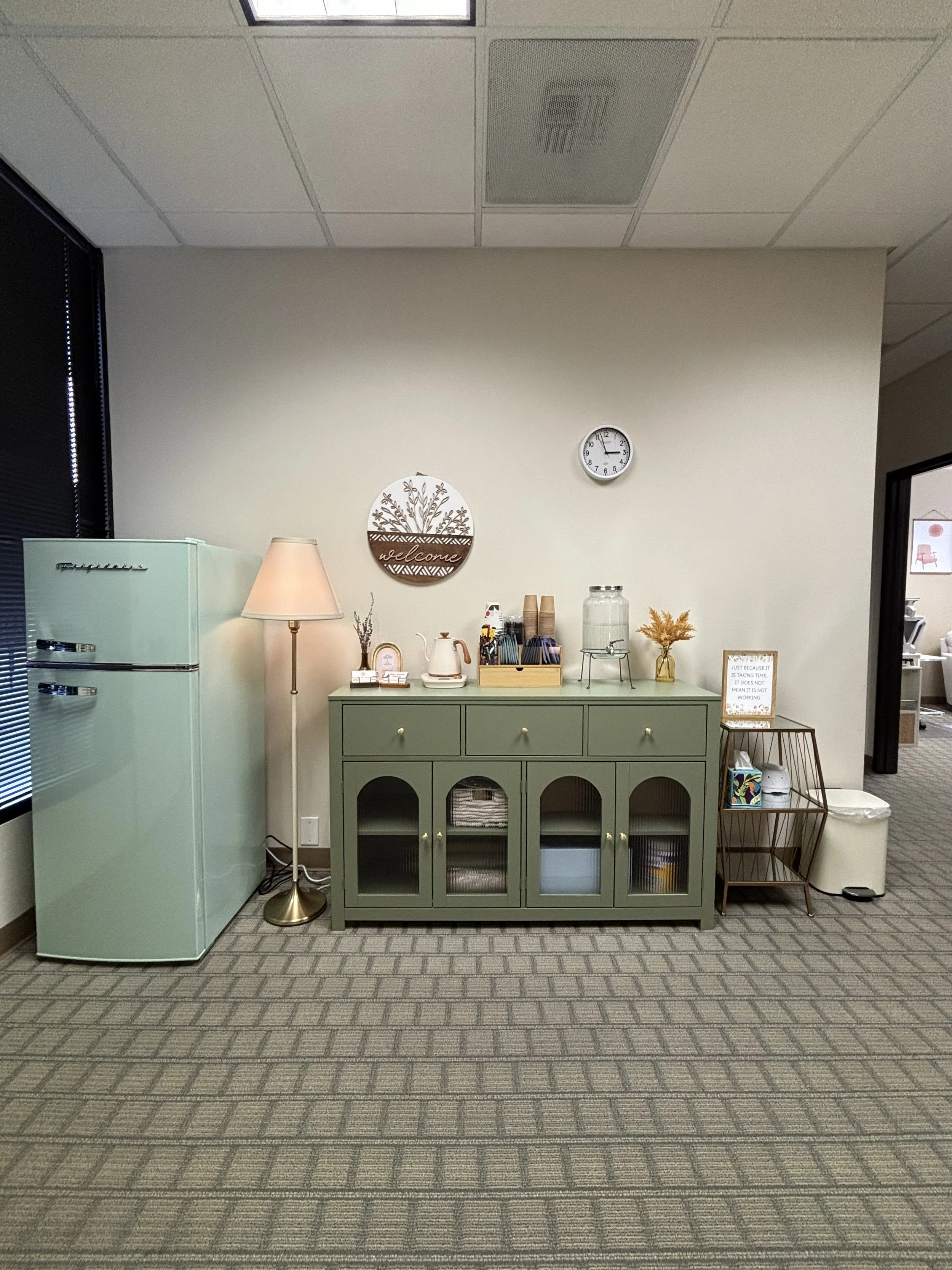 A room corner with a mint green refrigerator, a green sideboard decorated with a table lamp, a small clock, and various decor items. The floor is carpeted with a patterned gray carpet, and the room has a beige wall with a "welcome" sign and a round wall clock showing 1:01. To the right is a small table with a framed quote, and a wire basket with books and a tissue box. The ceiling is white with square tiles and visible light fixtures.