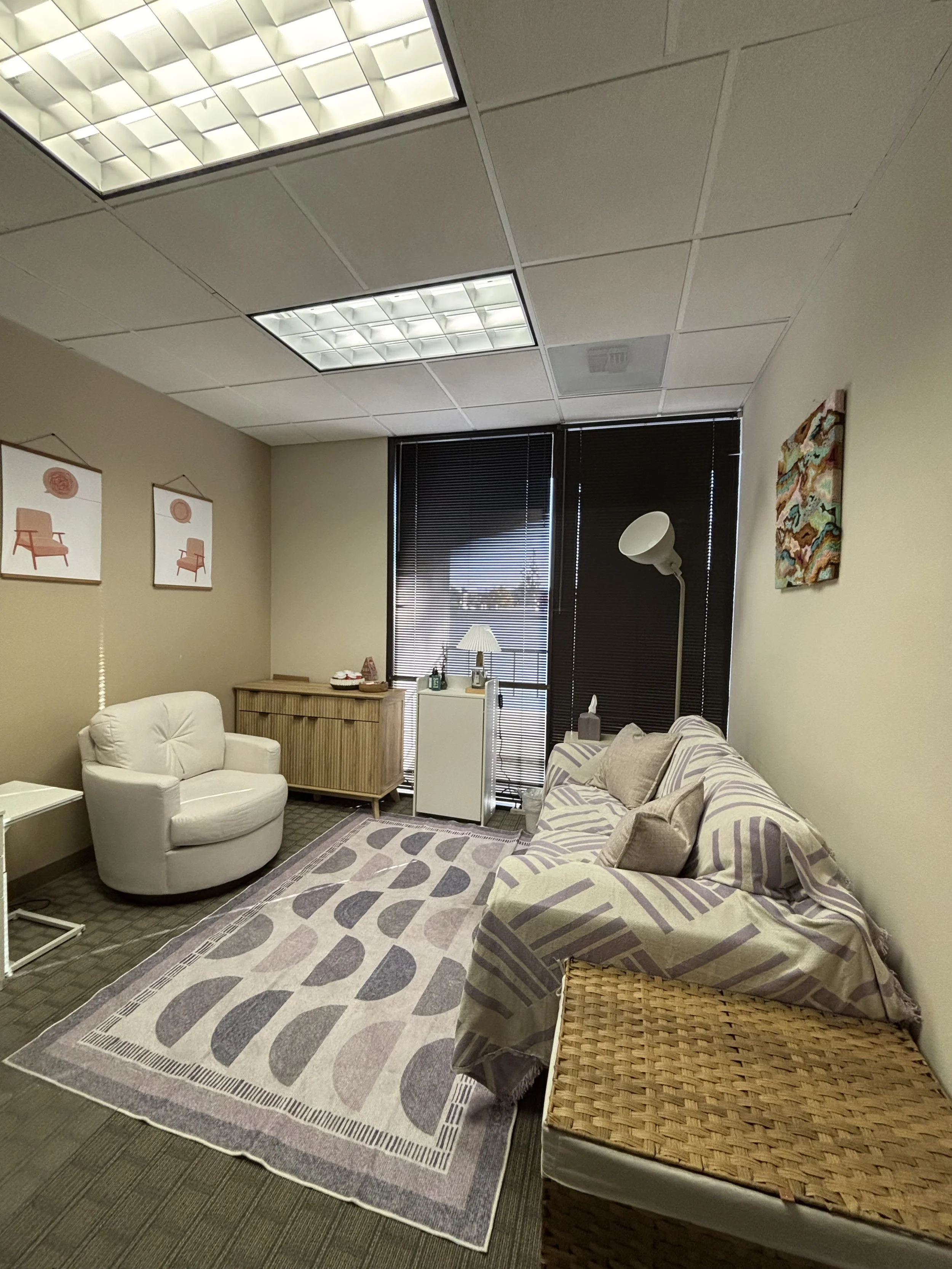 A cozy office or lounge space with beige walls and a gray patterned rug. There is a white armchair, a sofa with purple and beige striped cover, and a light wood cabinet with a lamp and decorative items. The room has large windows with black blinds, a tall white floor lamp, and artwork on the walls, including a colorful abstract piece on the right and two framed prints of chairs on the left. The ceiling has fluorescent lights.