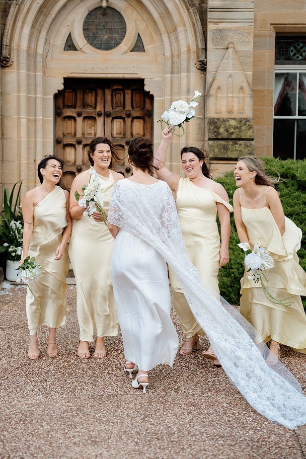 A bride in a white wedding dress with a lace veil dancing with five bridesmaids in matching cream-colored dresses outside a church with a large wooden door, smiling and holding bouquets of white flowers.
