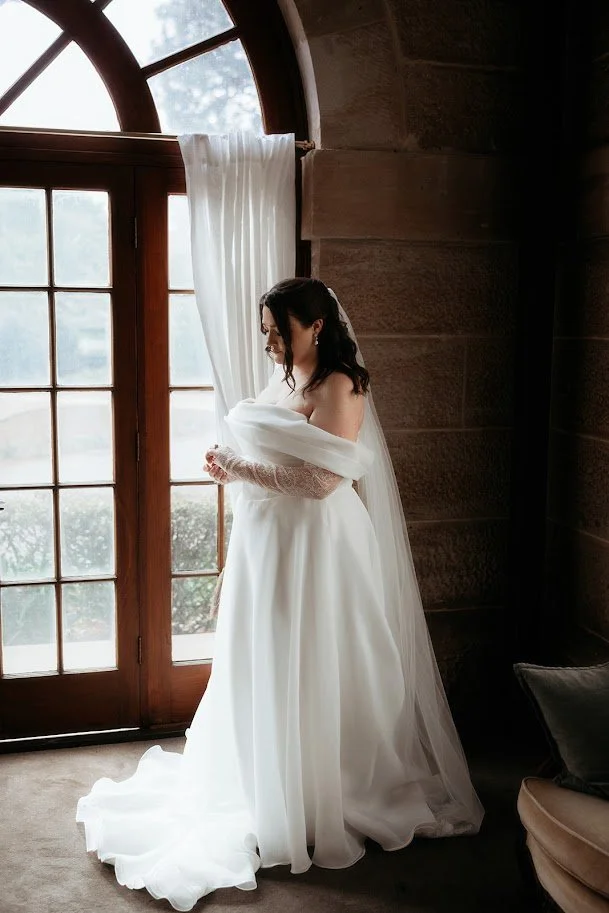 A bride by a tall timber window in a flowing off-the-shoulder white wedding gown with a long sheer veil and lace gloves, her steamed and finished dress falling smoothly to the floor. Wedding dress steaming, Toowoomba.