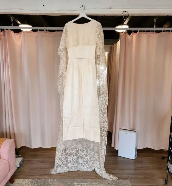 A wedding dress hanging on a white hanger in front of pink curtains, with a pink chair on the left and a white air purifier on the right.