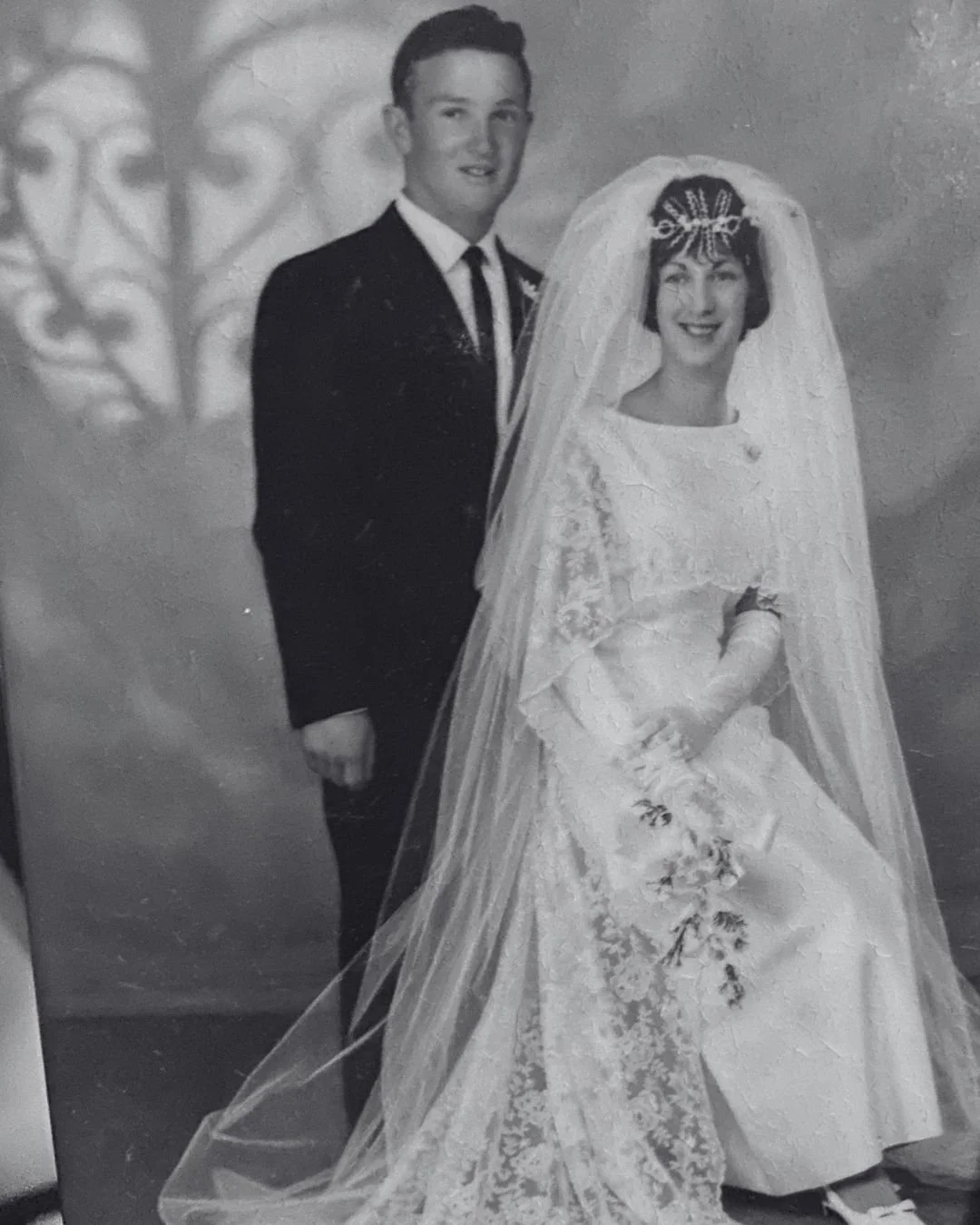 A black-and-white wedding photograph of a couple, with the bride wearing a lace wedding gown and veil, and the groom in a black suit, standing together and smiling.