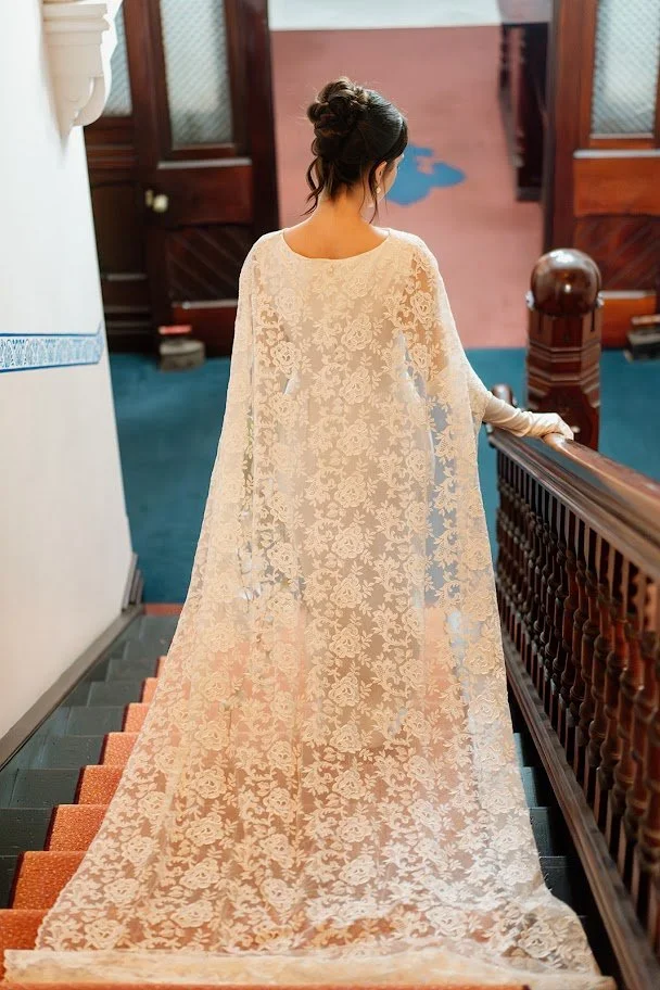 Woman in a long, cream-colored lace dress with an intricate floral pattern, standing on a staircase and facing away.