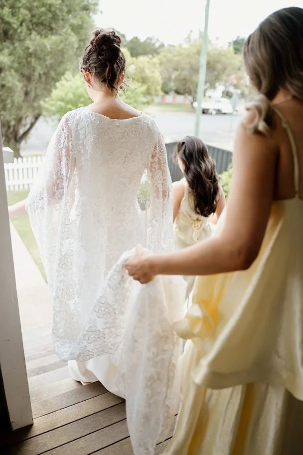 A bride in a lace wedding dress holding the train of her gown, with a young girl in a yellow dress helping her, and another woman in a yellow dress standing nearby, on a porch with a white picket fence in the background.