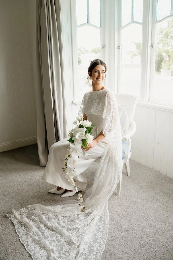 Bride sitting on a chair near a window, holding a bouquet, wearing a white wedding gown with lace details, and a long lace train on the floor.