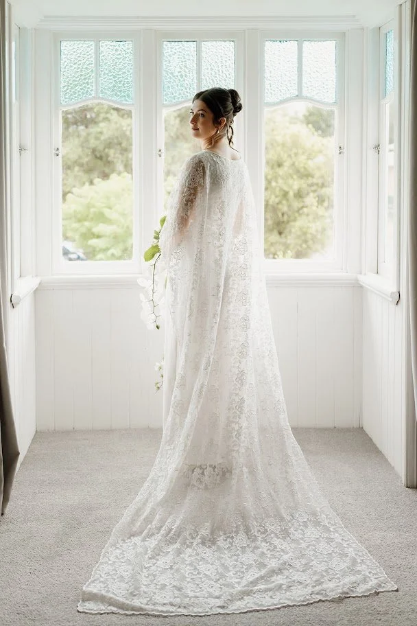 A woman in a white lace wedding dress standing by a window with a view of green trees outside, holding a white flower.