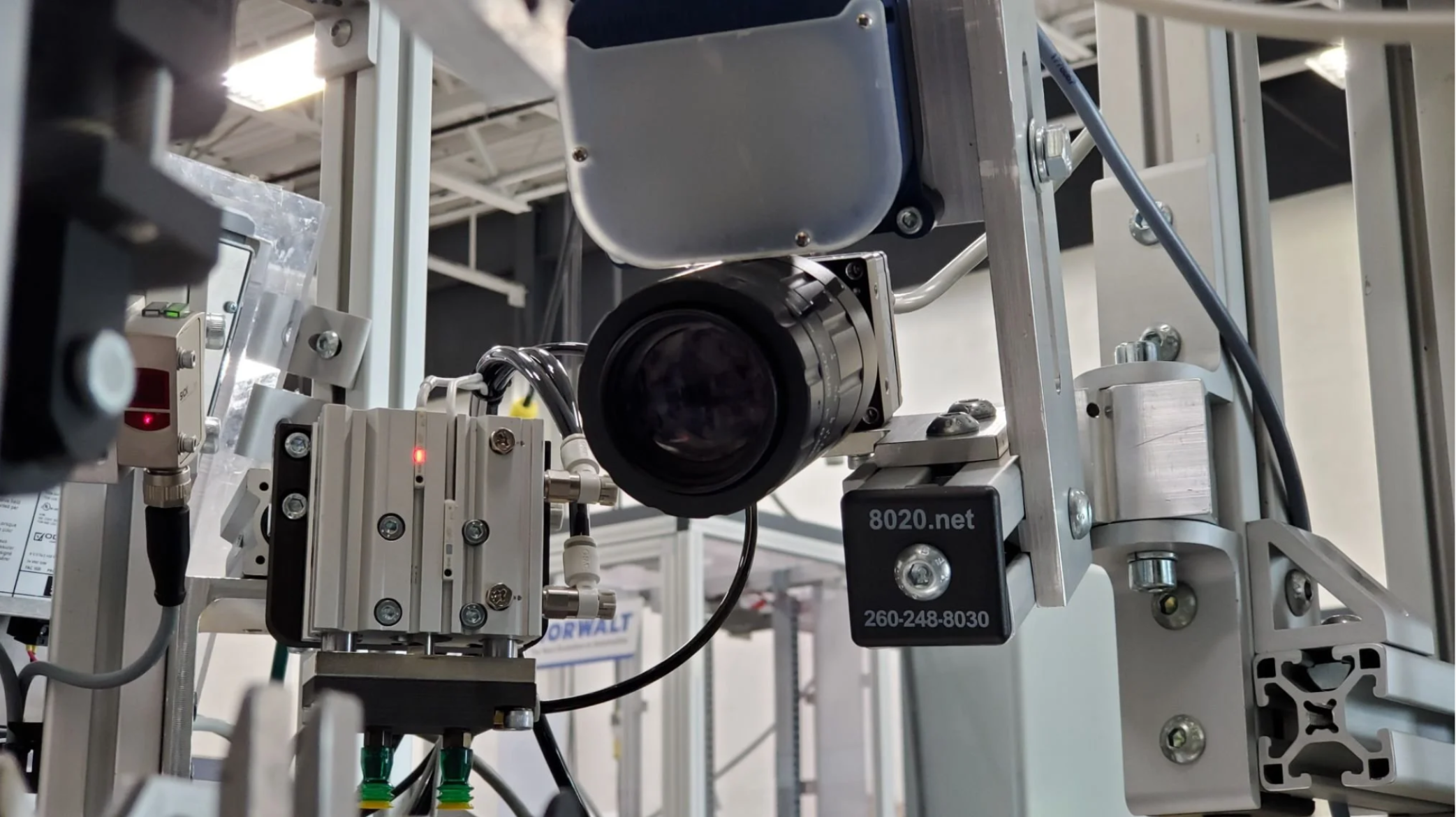 Close-up of industrial machinery with various metal components, wires, and a black camera or sensor in a factory setting. Line scan camera, bar lights.