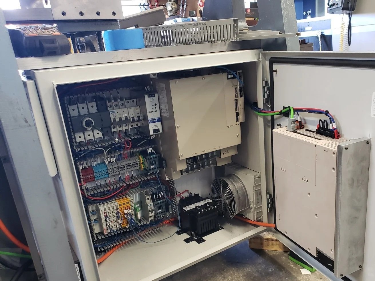Open electrical cabinet with various components, wires, and a cooling fan inside an industrial setting.