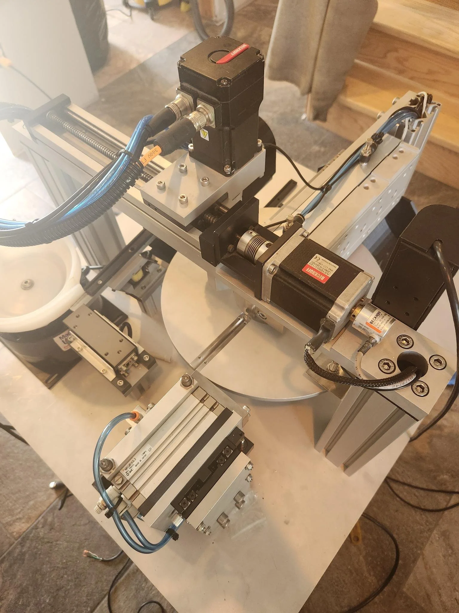 Close-up of a mechanical or industrial machine with various components, wires, and metal parts, situated on a white surface with a wooden and concrete background.