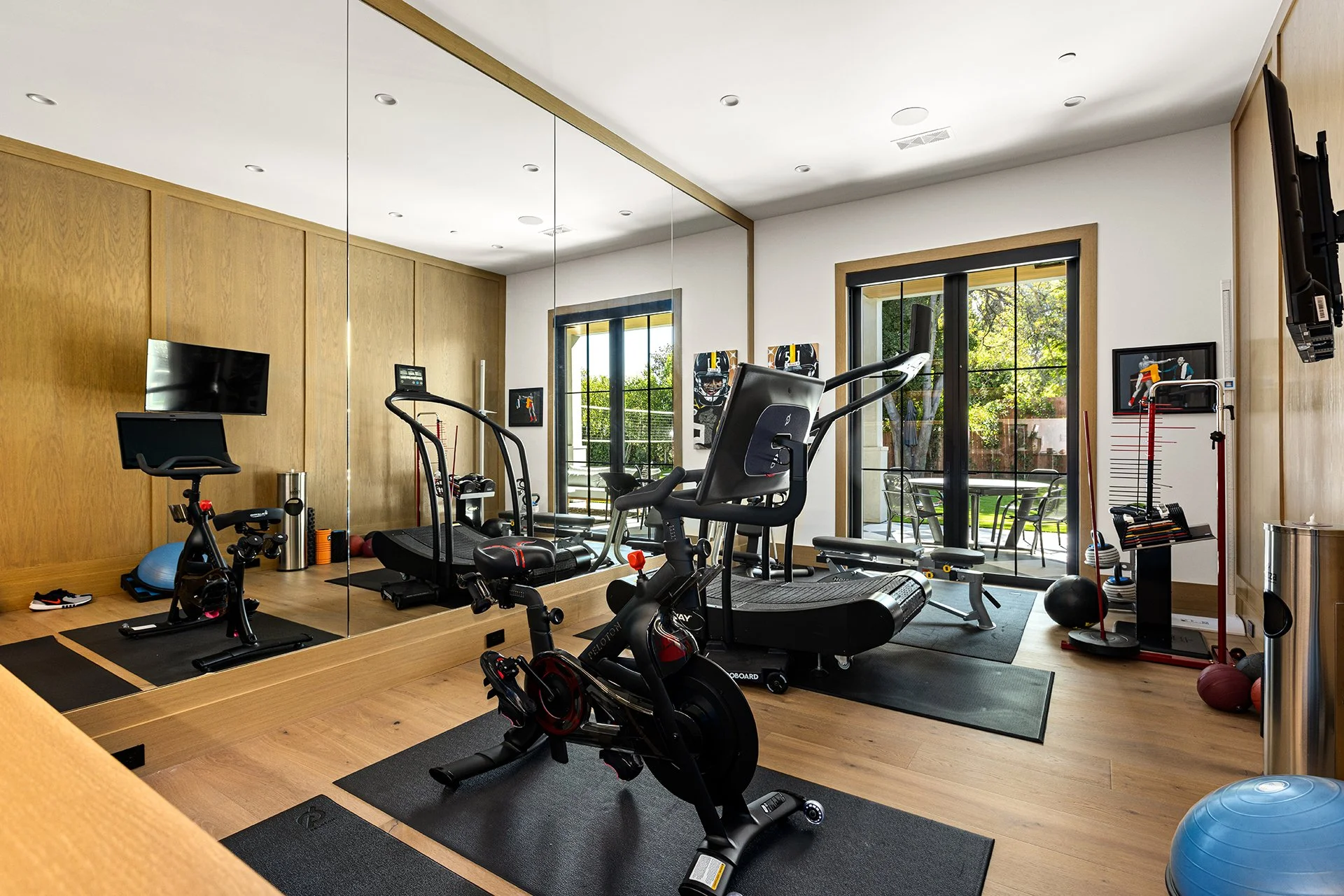 Home gym with exercise equipment including a stationary bike, treadmill, elliptical machine, weights, and a TV, with large windows overlooking a backyard.