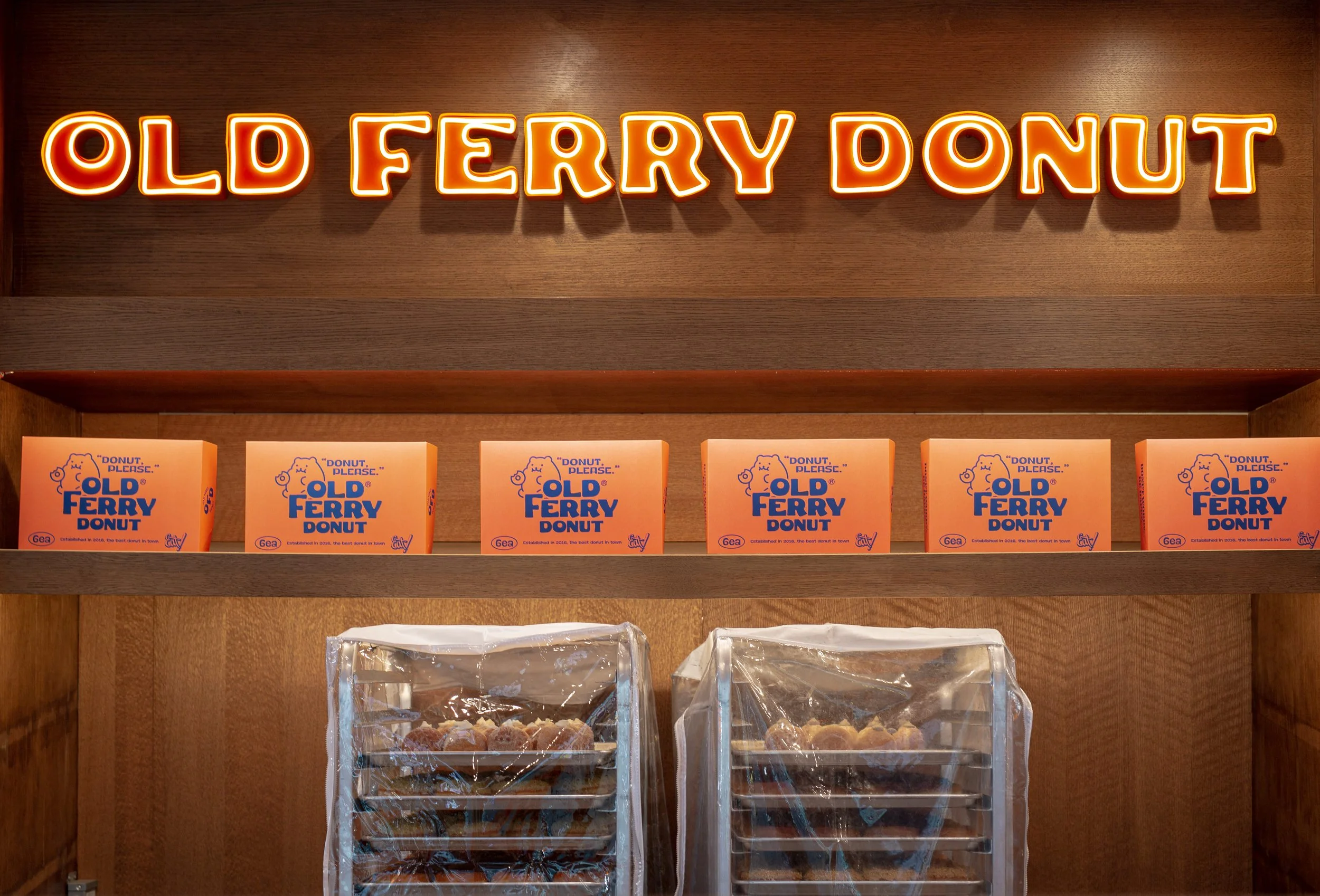 Shelf with boxes of Old Ferry Donut in front of a neon sign that reads 'Old Ferry Donut' in a wooden interior.