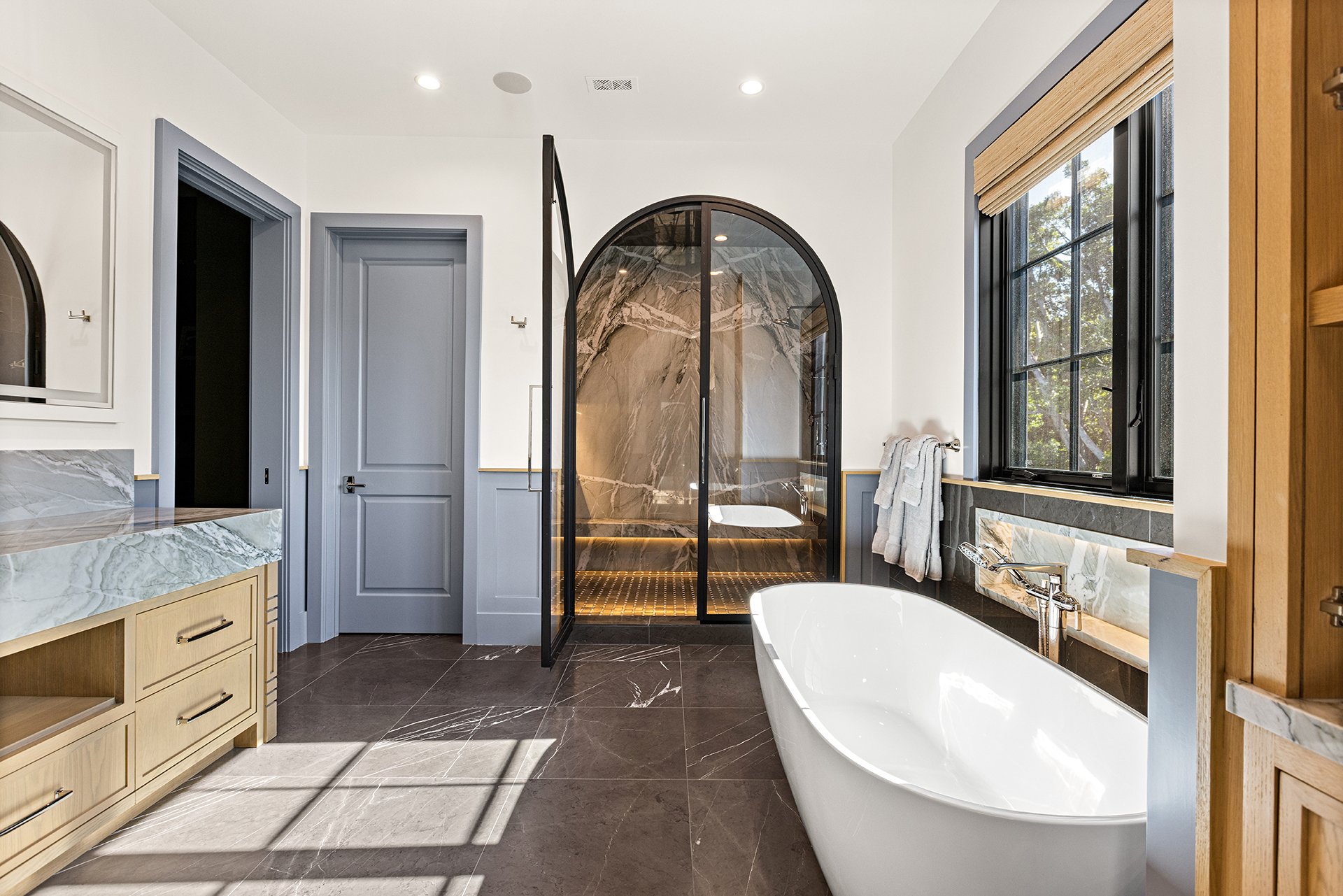 Modern bathroom with a white bathtub, a glass shower with marble walls, a vanity with a marble countertop, a large window with black framing, and gray doors.