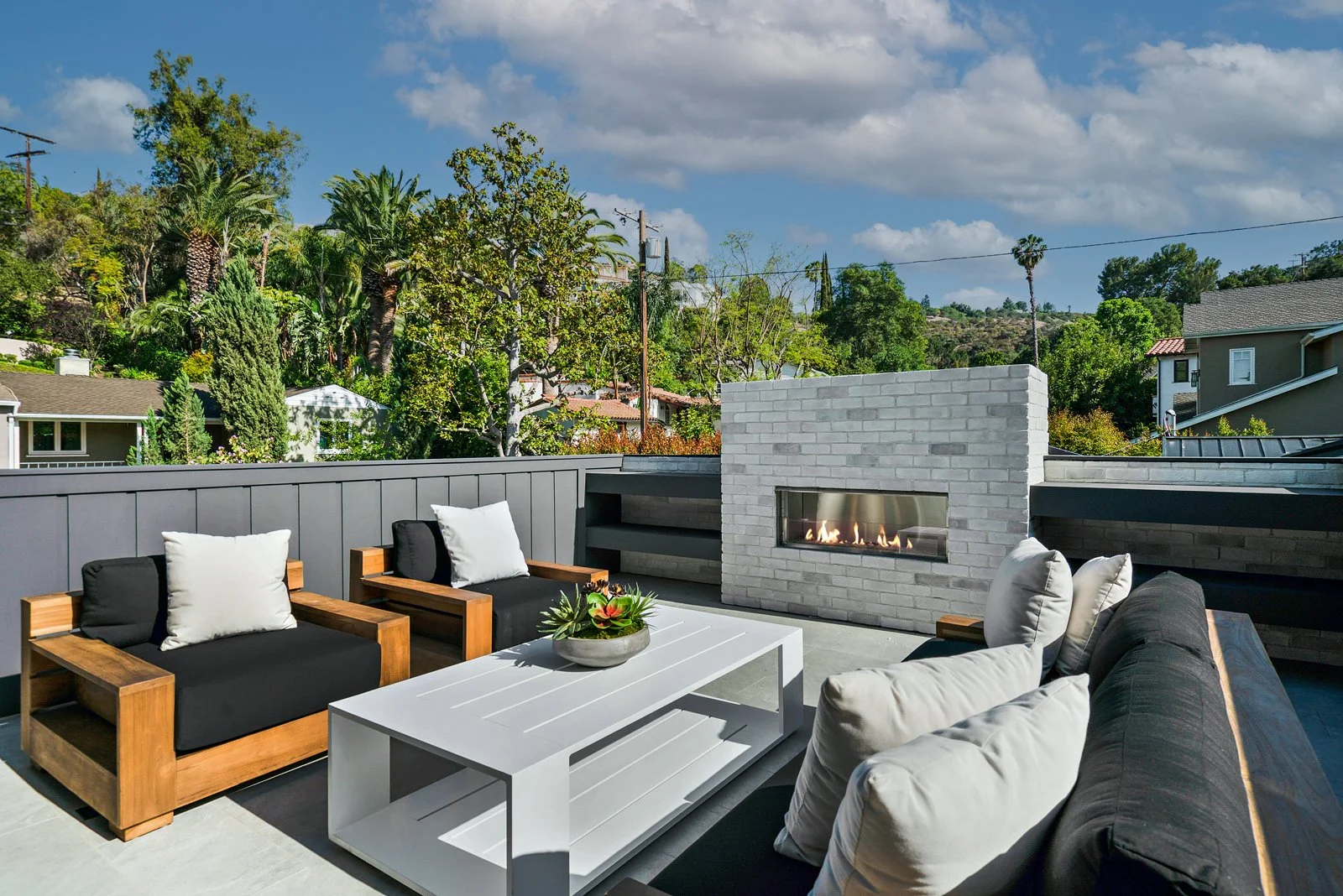 Outdoor patio with modern furniture, including wooden armchairs with black and white cushions, a white coffee table with a plant centerpiece, and a gray brick outdoor fireplace. Surrounded by lush greenery and trees under a partly cloudy sky.