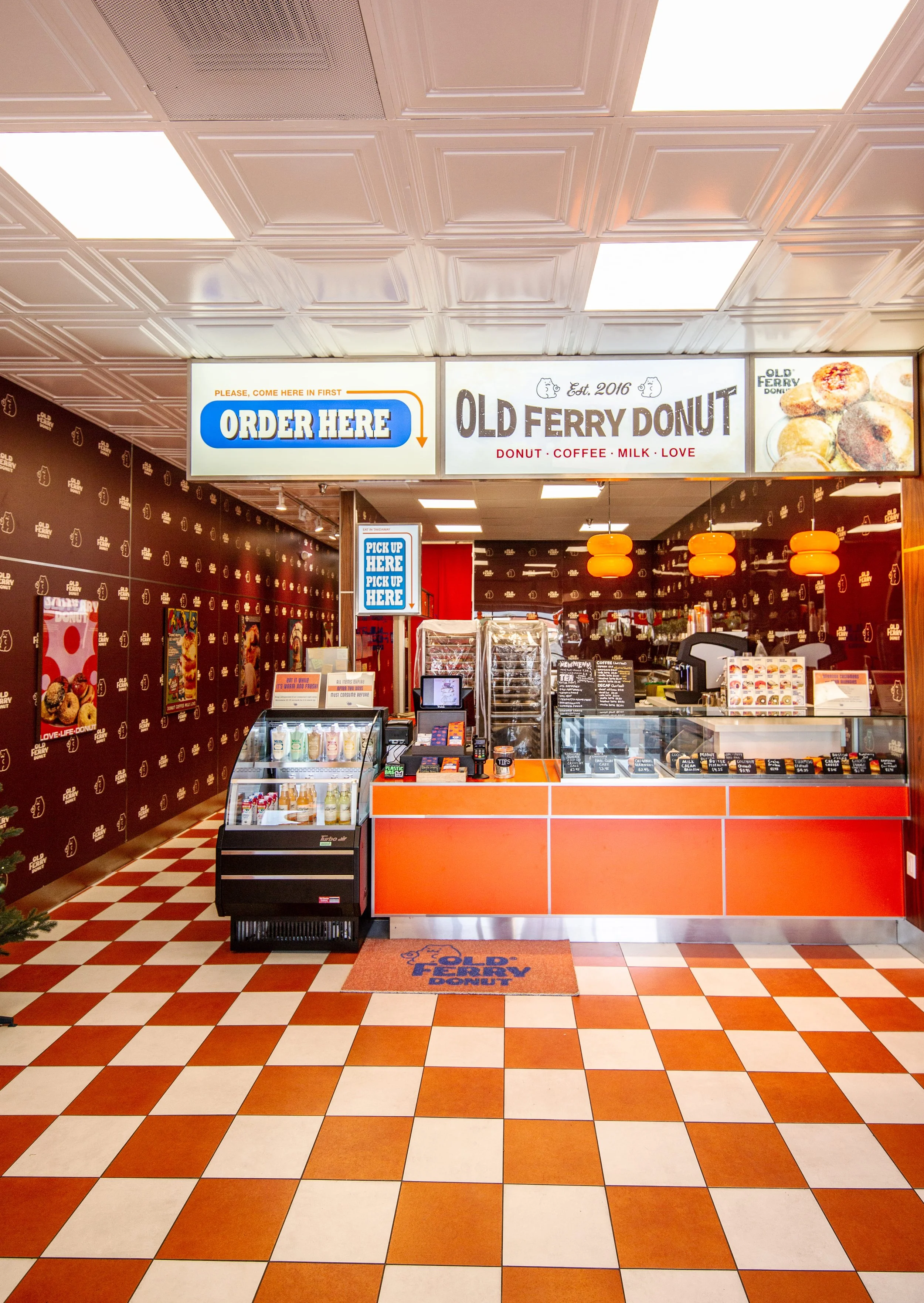 Fast food counter for Old Ferry Donut, offering donuts, coffee, milk, and other products with signage indicating where to order and pick up, and orange hanging lights.