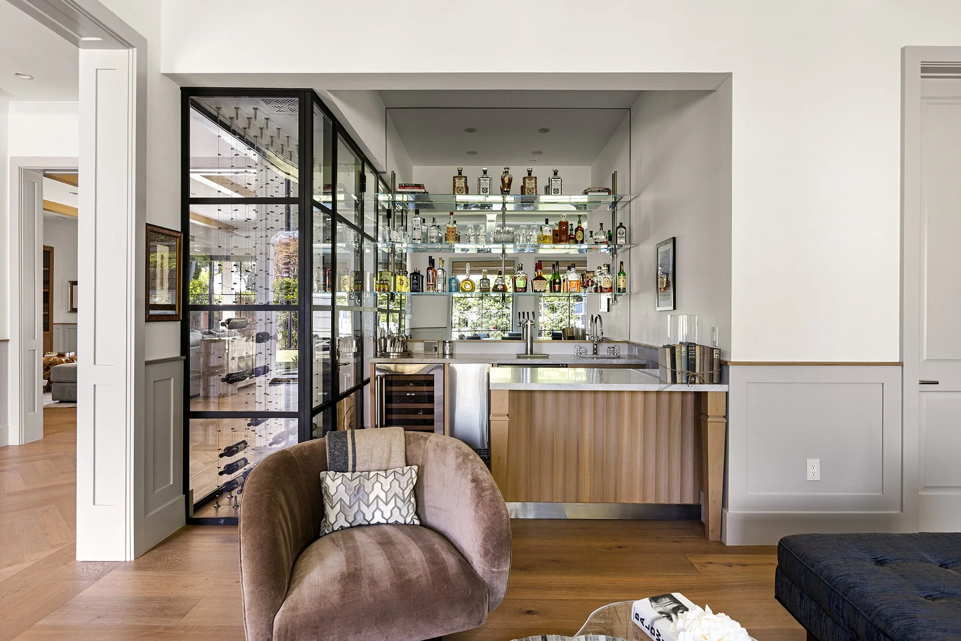 Living room with a white bar area featuring glass shelves stocked with various liquor bottles, a black metal and glass partition, and a brown velvet armchair with a patterned pillow.