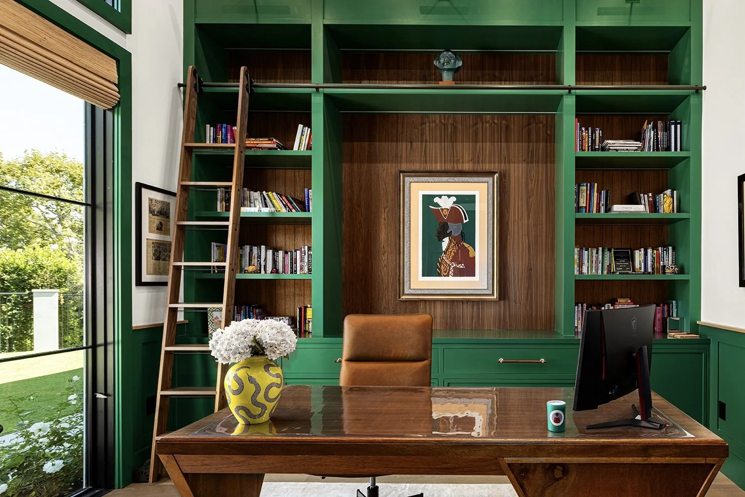 Modern home office with green built-in bookshelves, a wooden desk with a leather chair, a window with a view outside, a yellow decorative vase with white flowers, framed artwork, and a computer monitor.