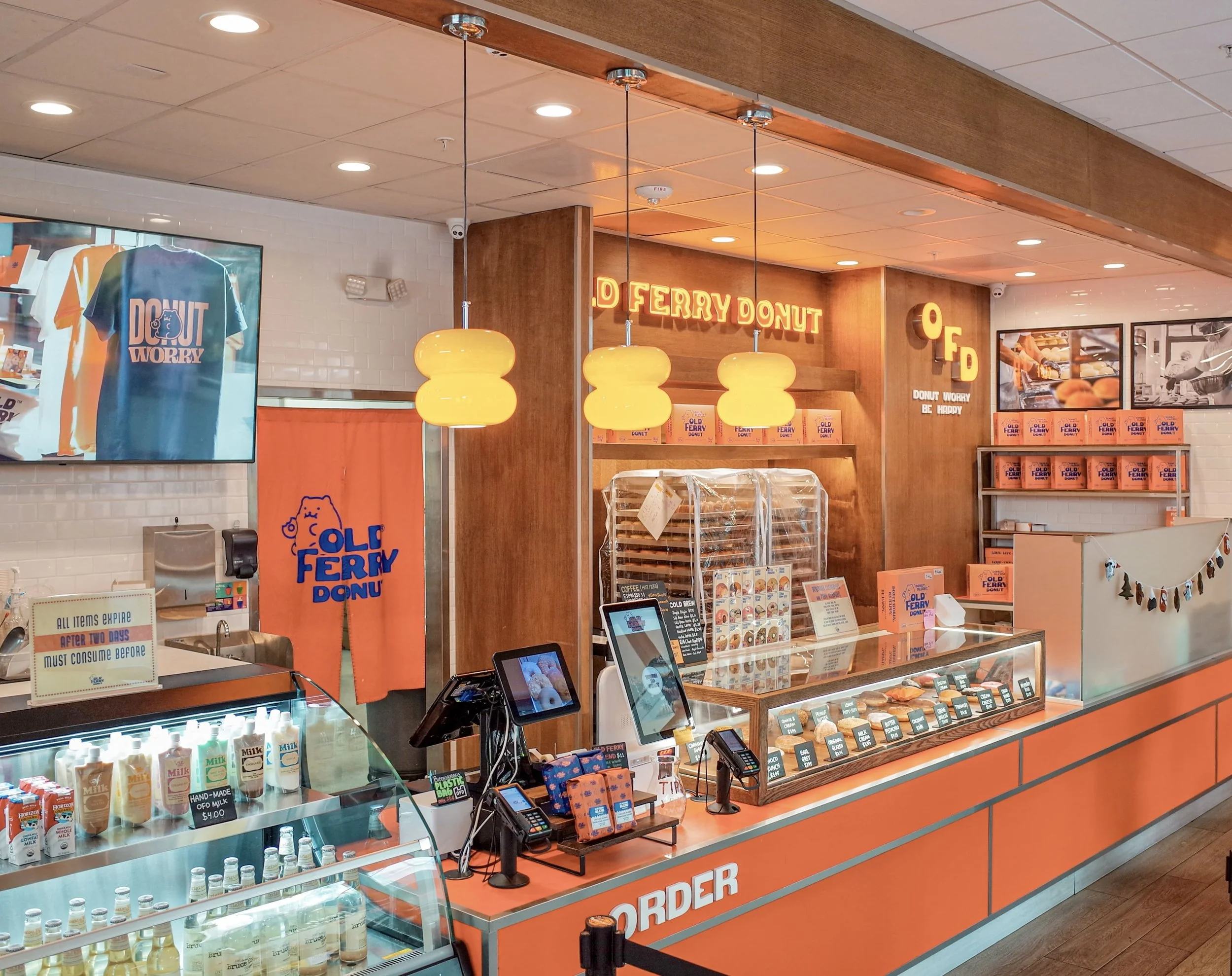 Interior of a donut shop called Old Ferry Donut, with orange and white decor, hanging yellow pendant lights, a display case with donuts, a digital menu, and promotional signs.