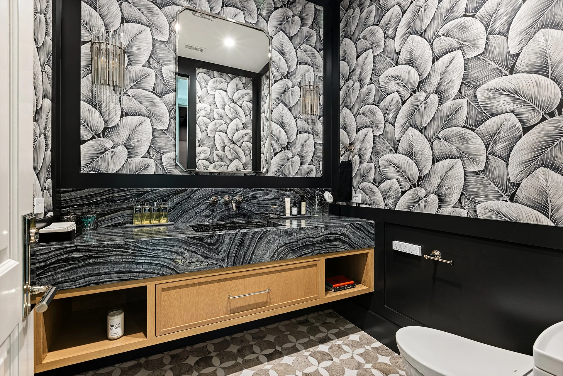Black and white leaf patterned wallpaper in a modern bathroom with a black-framed mirror, marbled black and gray countertop, wooden storage drawer, and a white toilet.