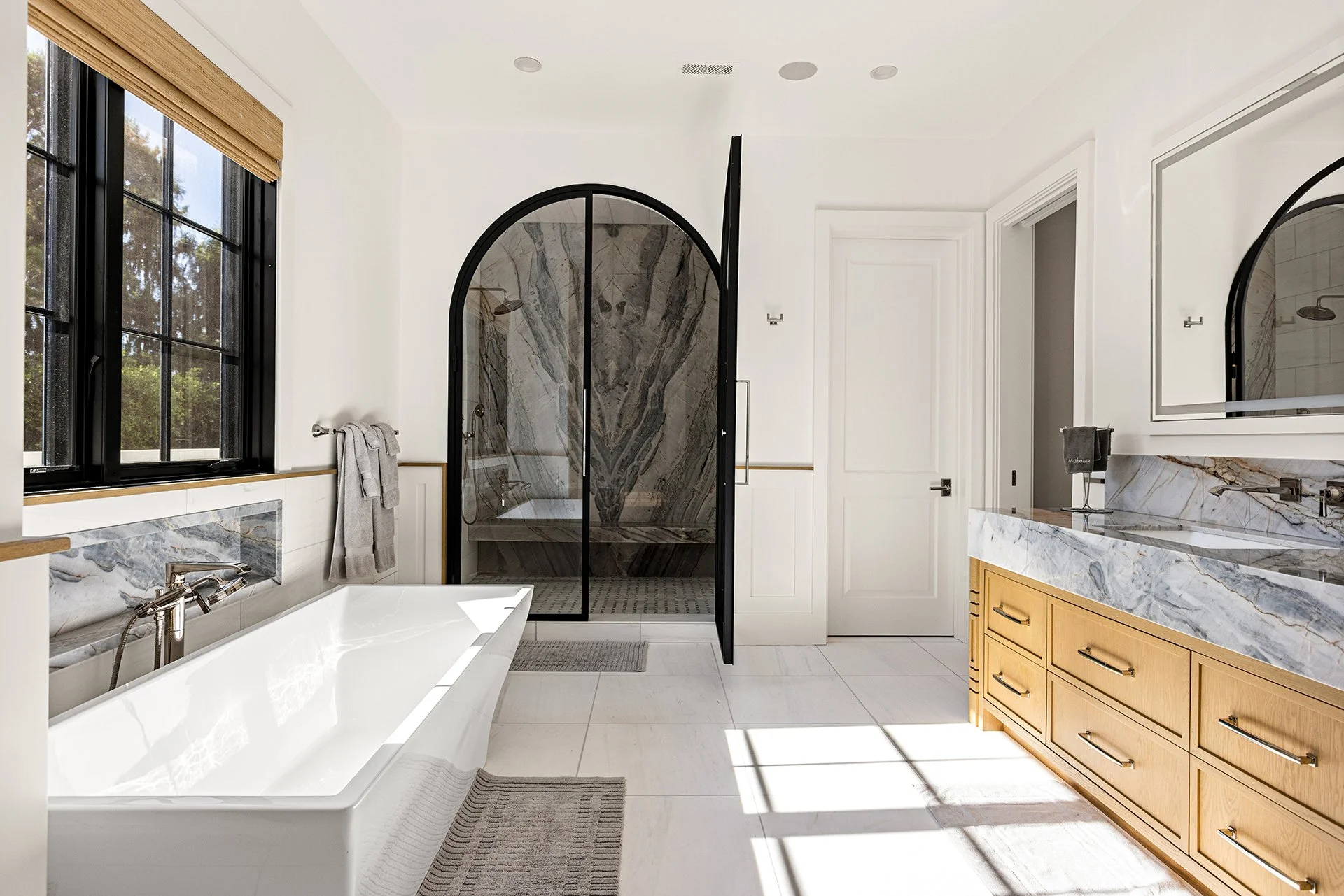 Modern bathroom with a white bathtub, a black-framed walk-in shower with marble walls, a wooden vanity with marble countertop and integrated mirror, large windows with black frames, and natural light.
