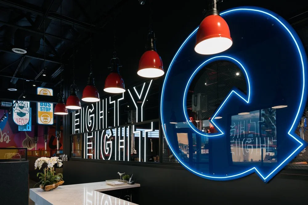 Exterior of a modern cafe with outdoor seating area, pendant lights, and a sign reading 'Mulholland' on the front. The white building has large windows and a landscaped sidewalk.