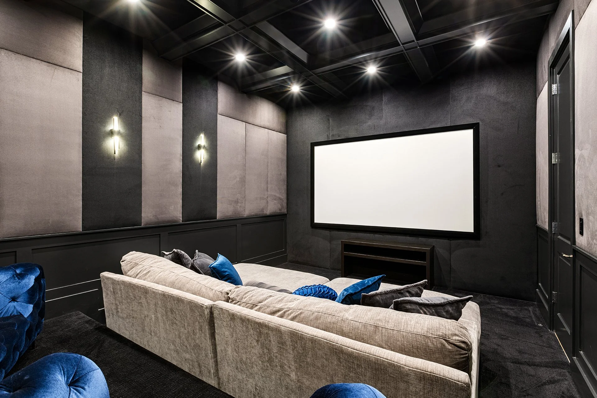 A modern home theater room with a large white screen, a gray sofa with blue and gray pillows, black walls, and velvet blue chairs, dimly lit with ceiling and wall lights.