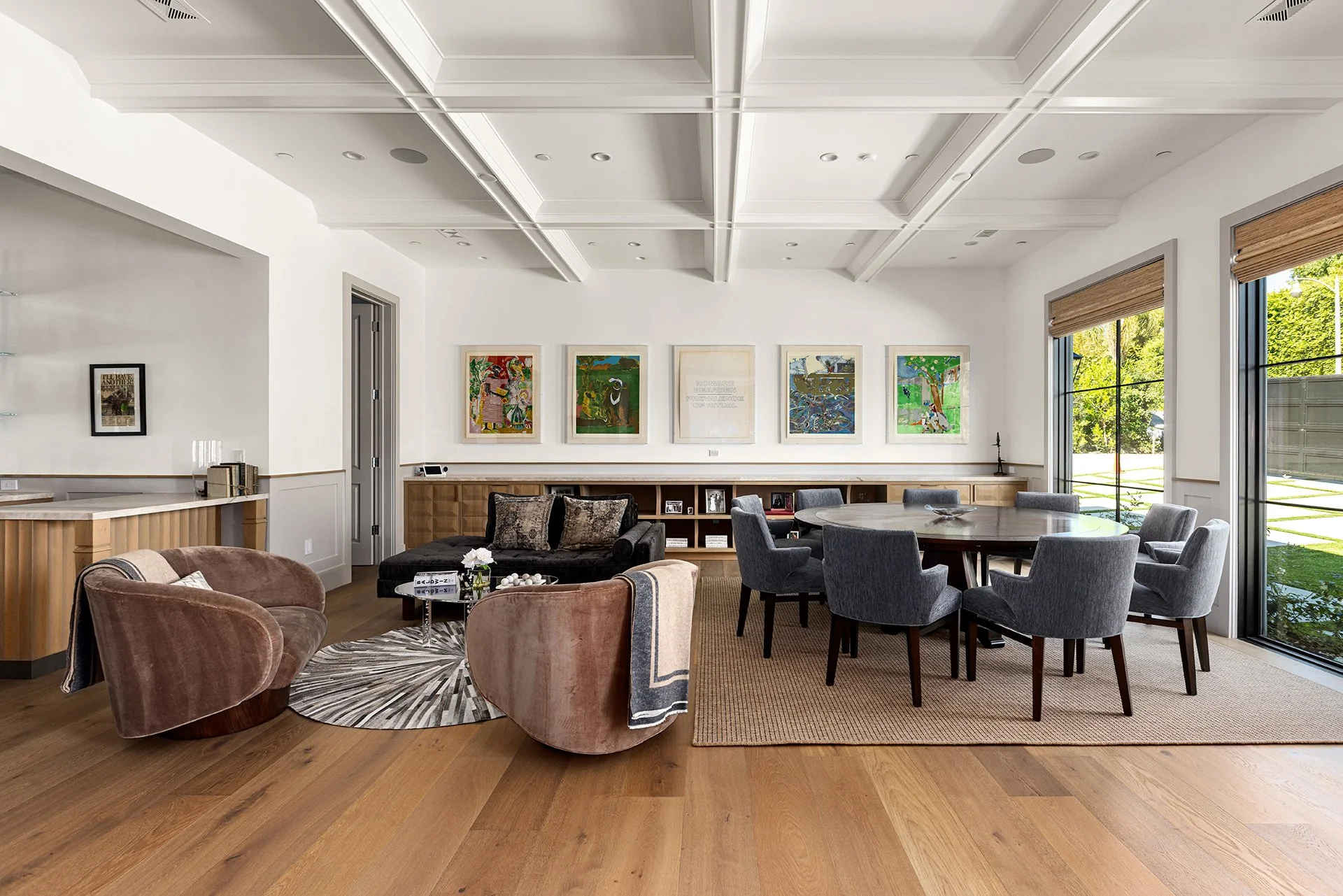 A modern living room with a white coffered ceiling, hardwood flooring, and a large window with blinds. The room features a black sofa, two pink velvet armchairs, and a round dining table with eight gray upholstered chairs. There are colorful painting