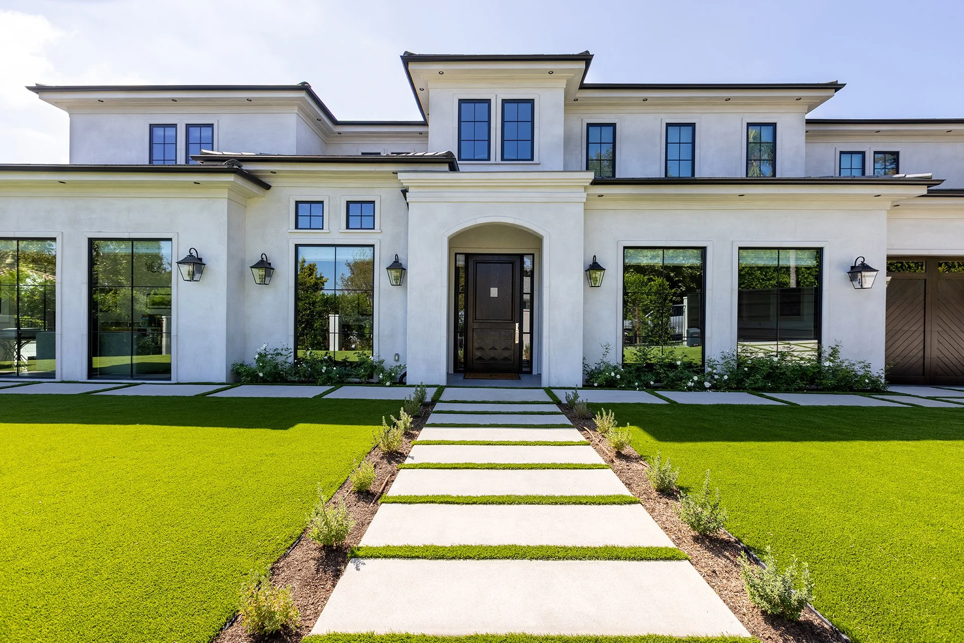 Front view of a modern white two-story house with large glass windows and a black front door, surrounded by a well-maintained lawn with a concrete pathway leading to the entrance.
