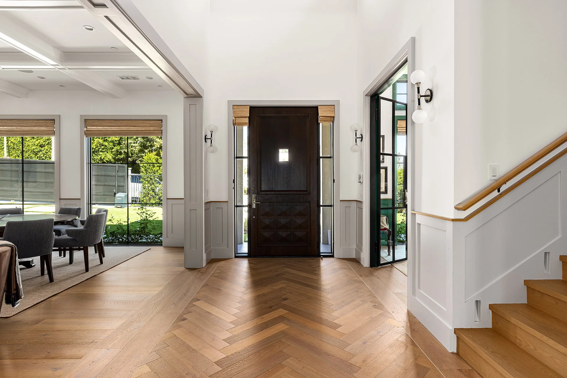 Entryway of a modern house with wooden flooring, a dark front door, and glass sidelights, adjacent to a dining area with windows and a patio outside.