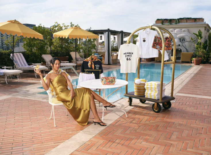 A woman in a gold dress sitting by a poolside at a clothing boutique, with a clothing rack holding shirts and accessories nearby, and pool chairs with umbrellas in the background.