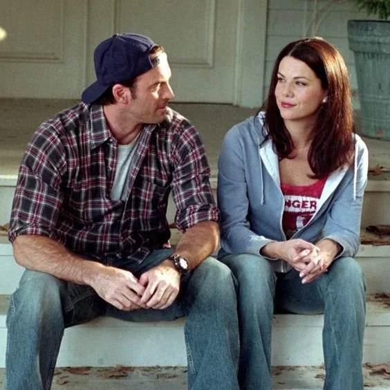 A man and woman sitting on stairs outdoors, looking at each other. The man is wearing a backward cap, plaid shirt, and jeans. The woman has red hair and is wearing a blue hoodie, red T-shirt, and jeans.