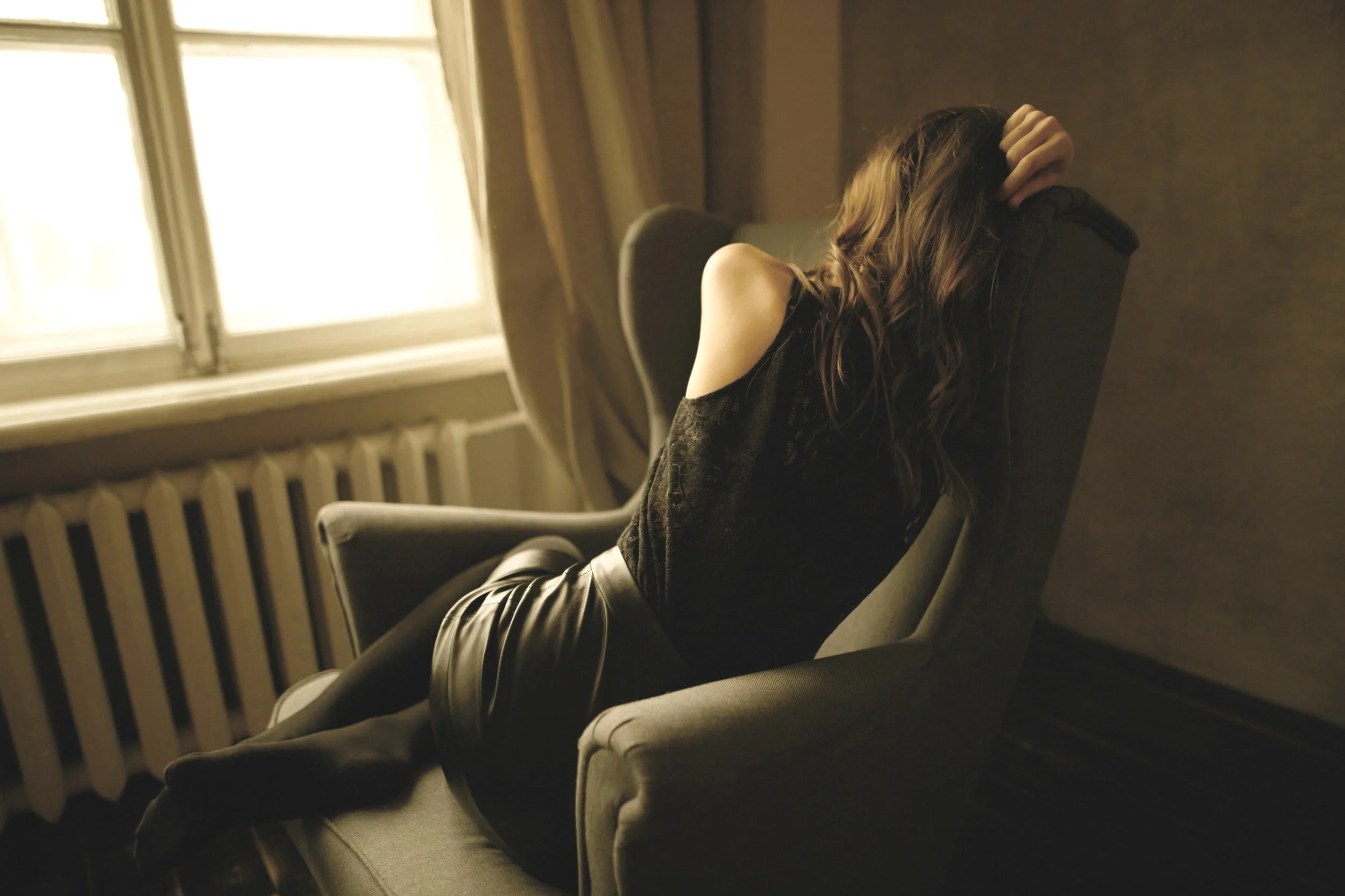 A woman with long wavy hair sitting in a dimly lit room on a gray armchair, near a window with sunlight streaming through light-colored curtains.