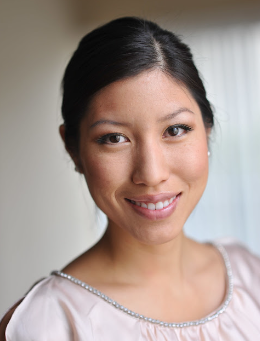 Portrait of a smiling woman with dark hair, wearing a light-colored blouse, standing indoors.