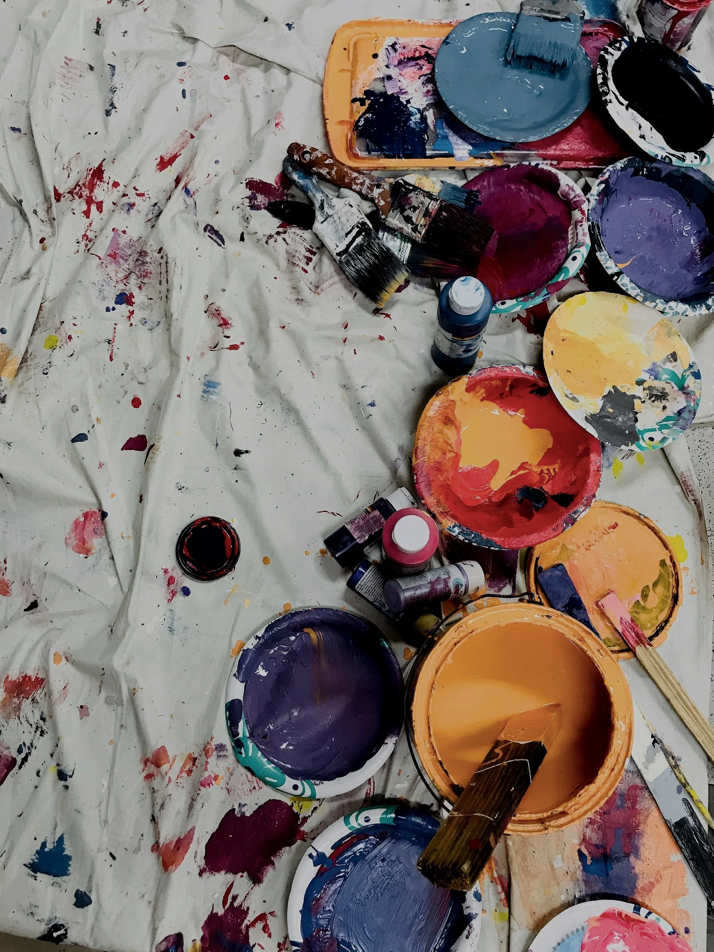 Assorted paint supplies on a splattered canvas, including various colored paint plates, brushes, and bottles.