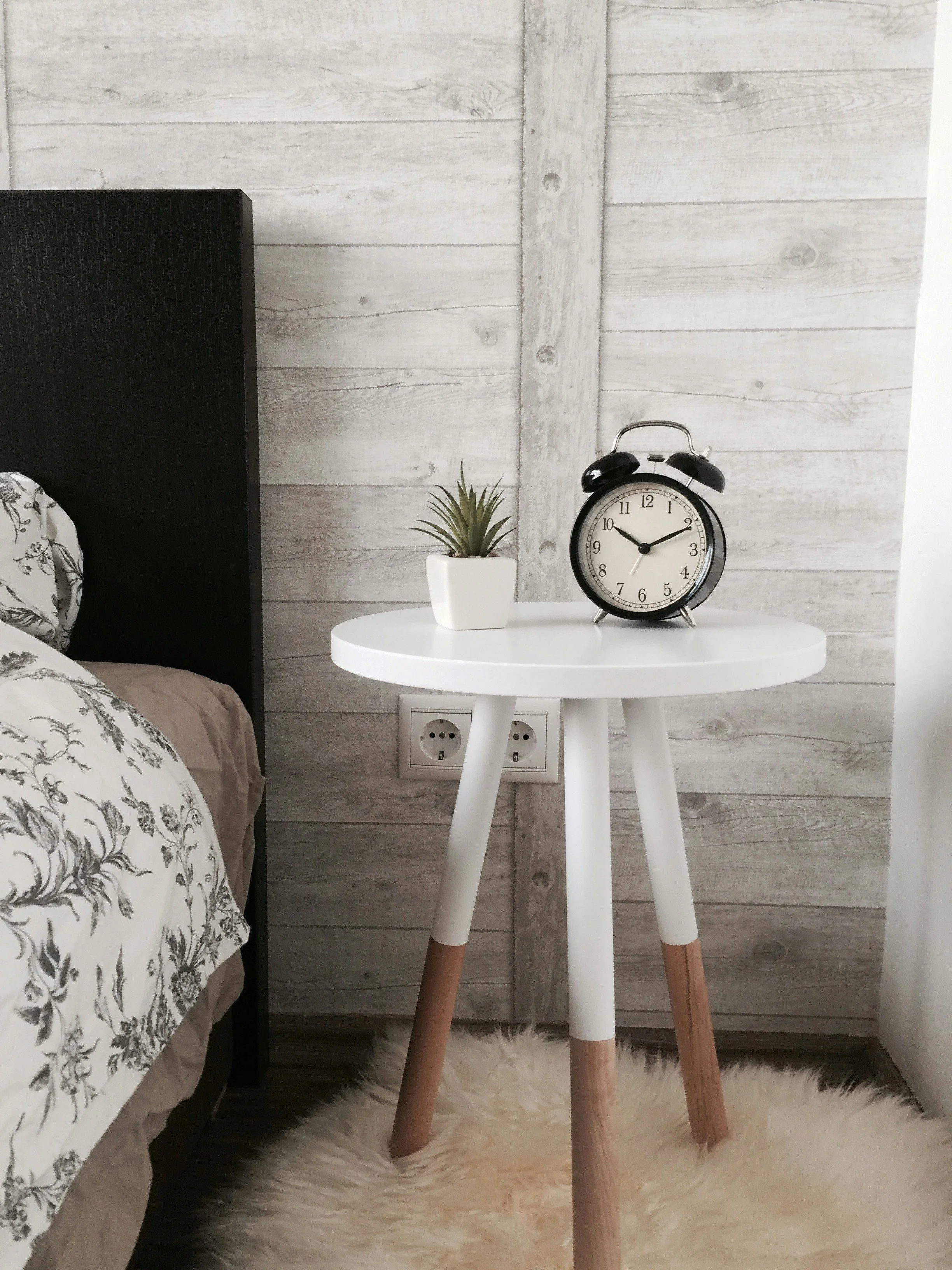 White round side table with wooden legs, holding a small potted plant and a retro black alarm clock, beside a bed with floral bedding, set against a gray wooden wall with electrical outlets.