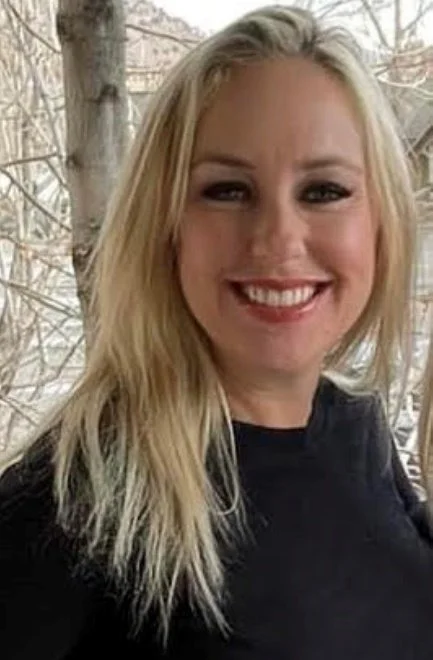 Smiling woman with long blonde hair wearing a black top, standing outdoors with trees in the background.
