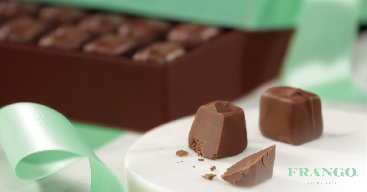 Chocolate truffles with a mint green ribbon, a brown box filled with chocolates in the background, and a white marble surface with chocolate pieces on it.