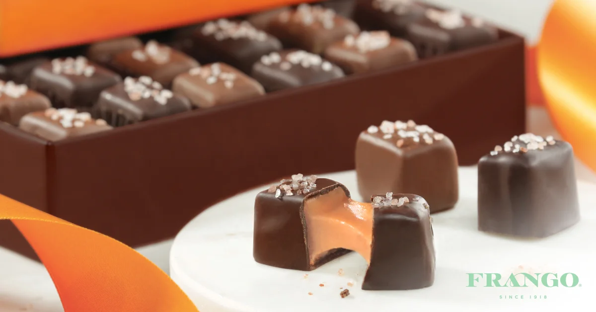 Assorted chocolates with sea salt on top, one cut open to show caramel filling, displayed on a white surface with an orange ribbon and a box in the background.