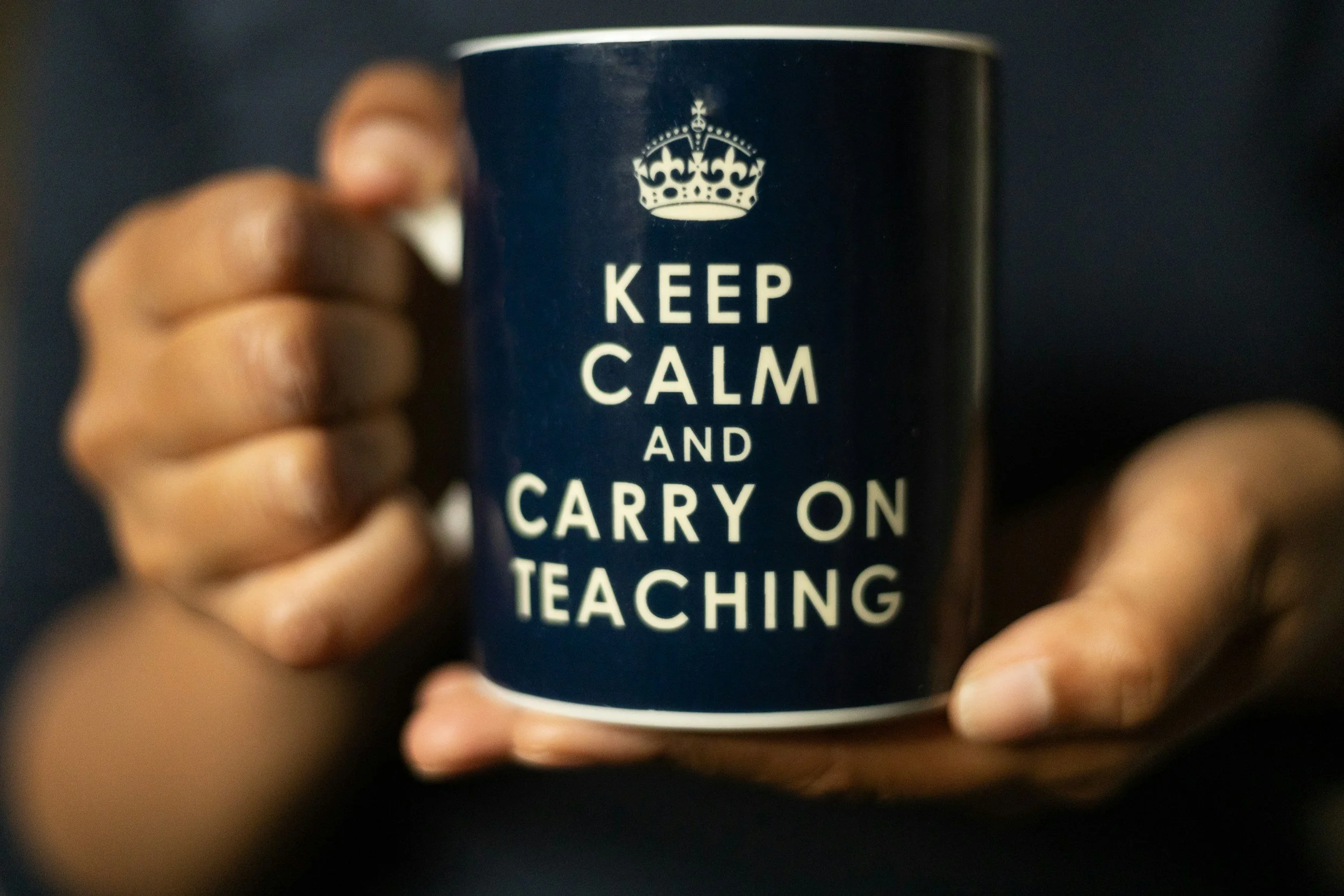Close-up of a dark-colored mug with the phrase "KEEP CALM AND CARRY ON TEACHING" in white text, being held by a person's hand.