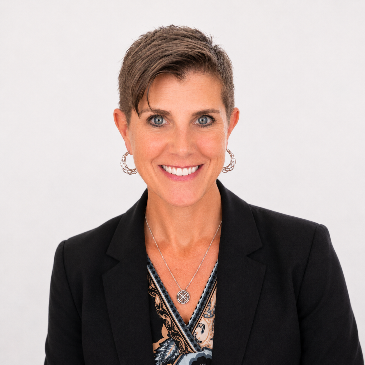 A woman with short brown hair, blue eyes, and a bright smile, wearing a black blazer, a patterned blouse, and silver jewelry, standing against a plain white background.