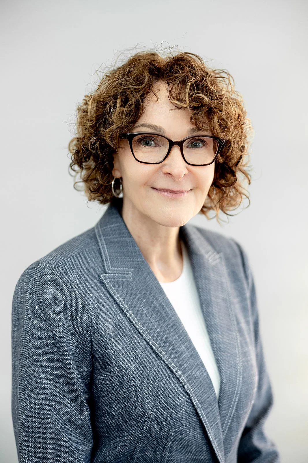 A woman with short curly hair wearing a white shirt and a blue blazer smiling in front of a plain, light-colored wall.
