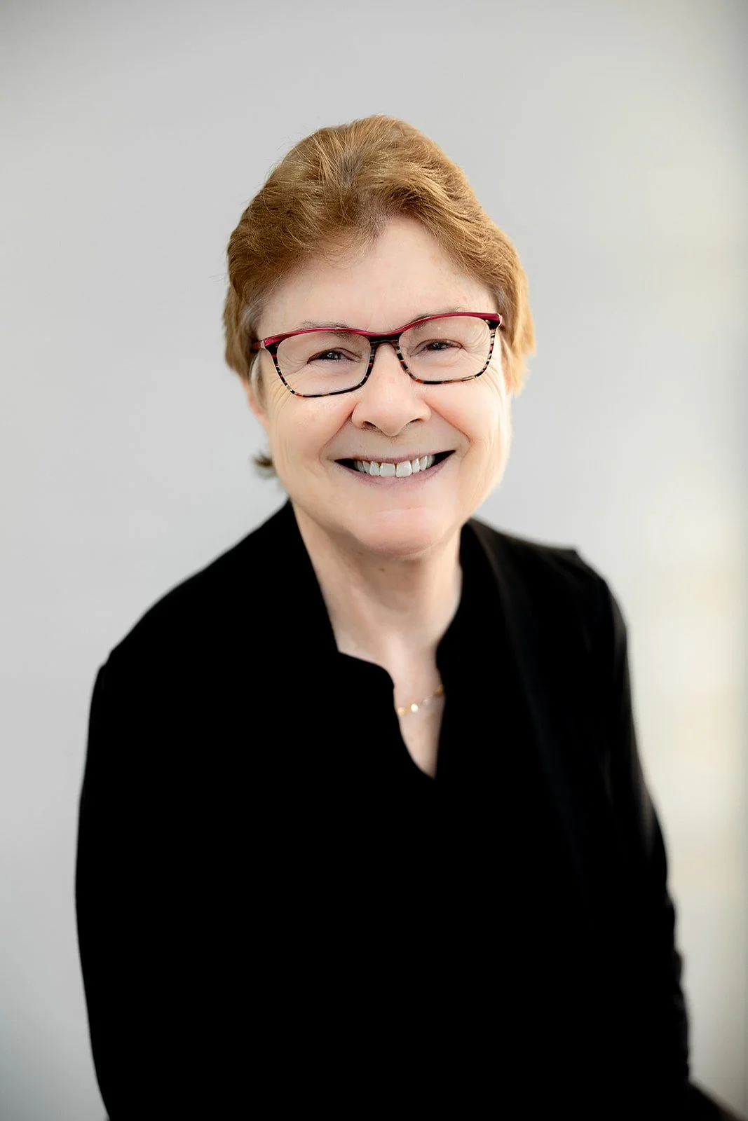 Portrait of an elderly woman with short reddish hair, wearing glasses, a red blazer, and a gold necklace, smiling against a plain light gray background.